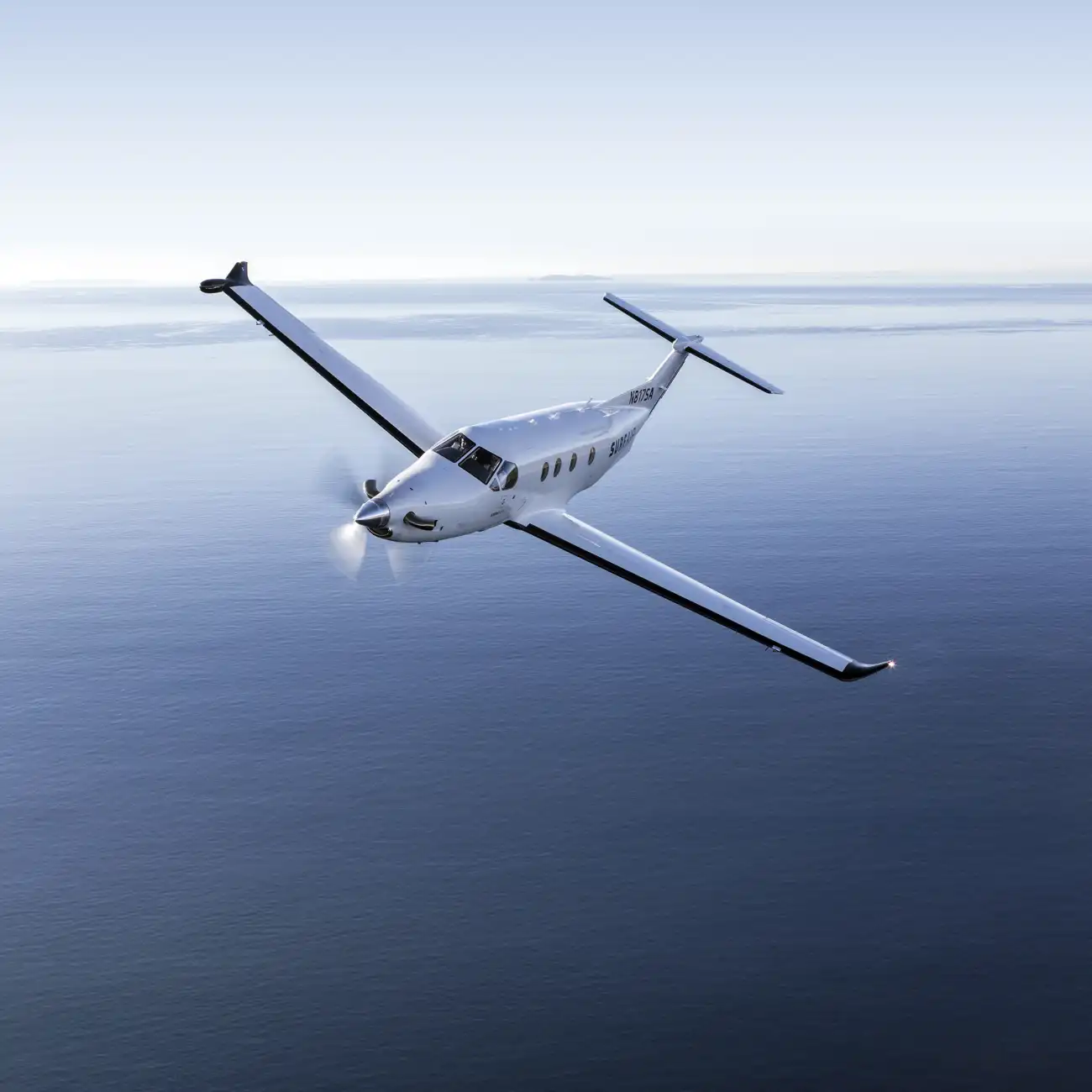 A gray-silver PC-12 flies over the water under a clear sky.