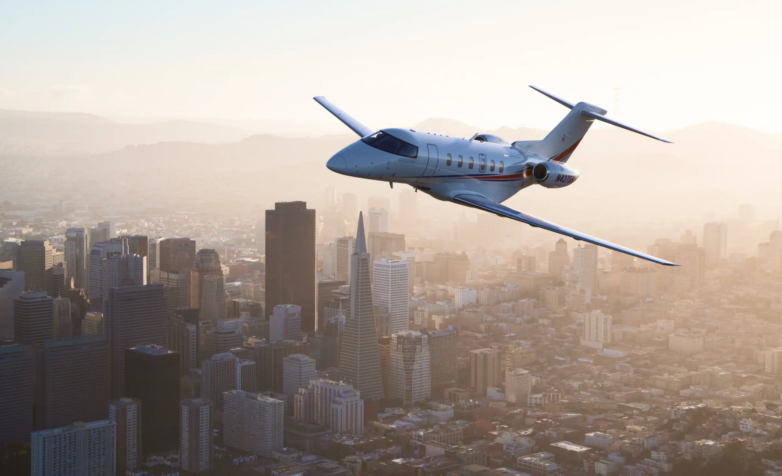 A PC-24 flies over a city with many high-rise buildings and skyscrapers, as the evening light casts warm hues across the sky.