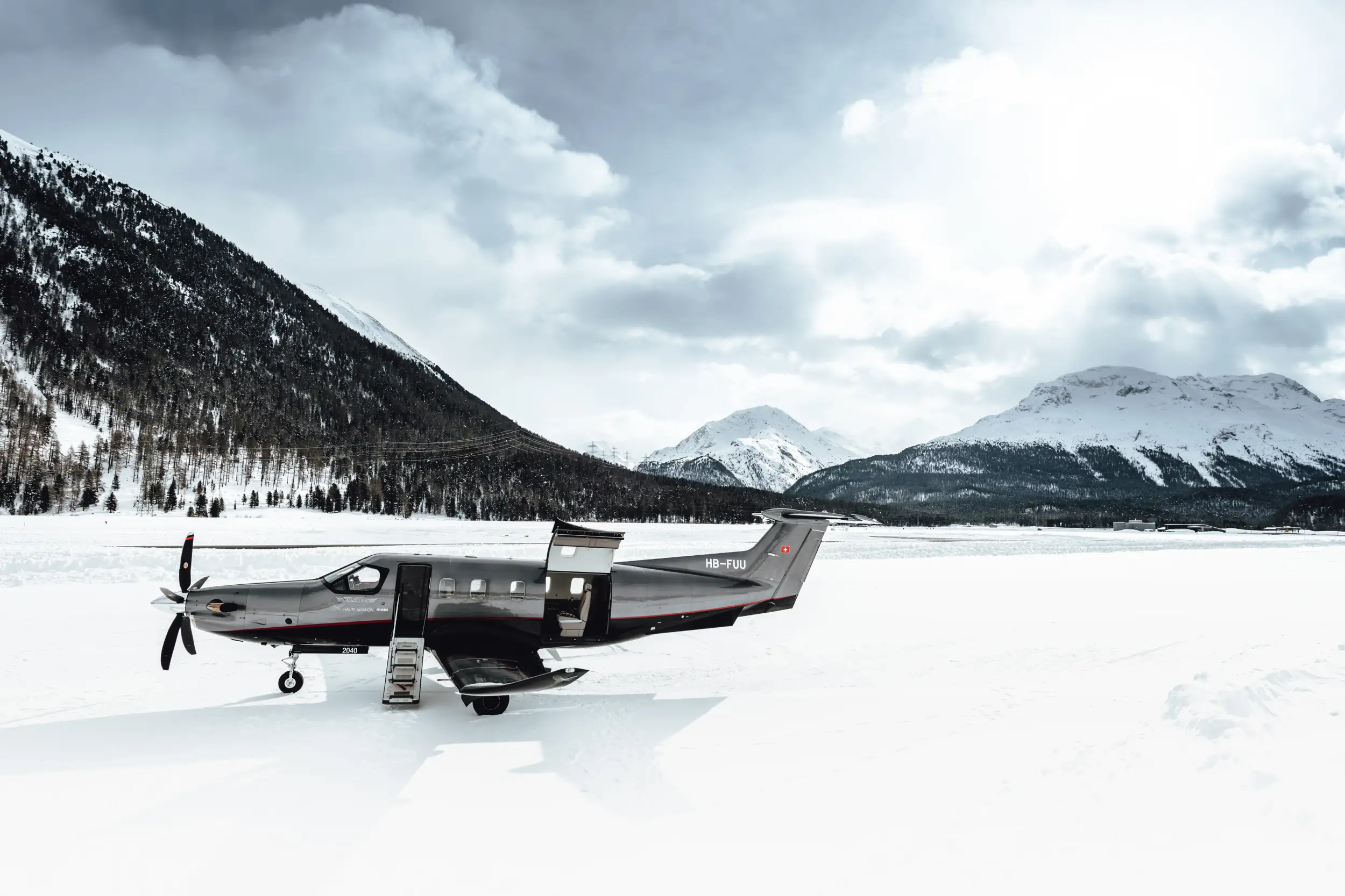 A PC-12 on snow, with open doors and mountains in the background.